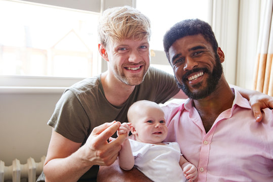 Portrait Of Loving Male Same Sex Couple Cuddling Baby Daughter On Sofa At Home Together