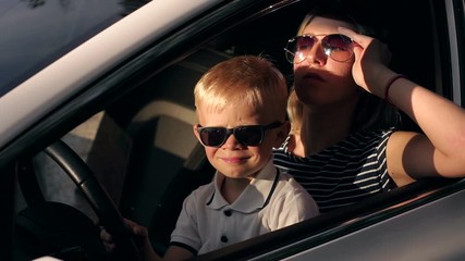 Portrait of a young mother with her son in sunglasses in the car, they sit together at the wheel, the bright sunset sun shines on their faces. Slow motion. Summer trip by car.