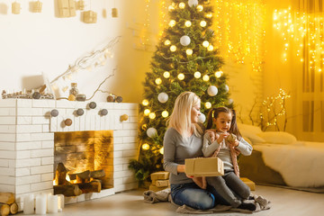 Young mother and her little daughter sitting by a fireplace in a cozy dark living room on Christmas eve