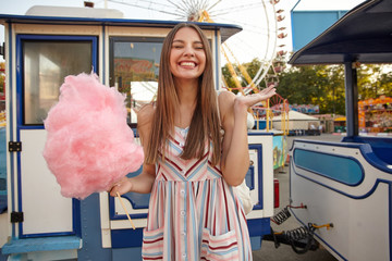 Positive young lovely brunette woman with long hair posing over amusement park, standing with pink cotton candy in hand and closed eyes, raising palm up and smiling cheerfully