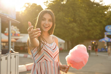 Sunny outdoor photo of charming brunette with brown hair posing over green park with pink cotton candy in hand, lending hand and inviting to walk with her, smiling happily to camera