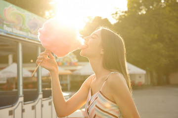 Outdoor sunny shot of happy pretty long haired lady in romantic dress posing over amusement park on warm summer day, eating pink cotton candy with big pleasure