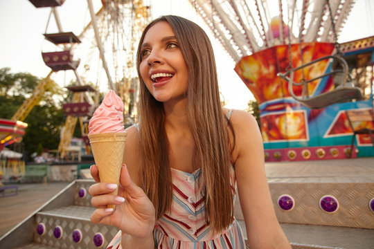 Portrait of happy brunette young lady posing outdoor on warm summer day with pink ice cream in hand, looking aside cheerfully, wearing light dress with straps