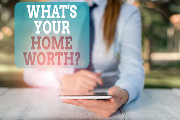 Writing note showing What S Your Home Worth Question. Business concept for Value of a house Property Cost Price Rate Female business person sitting by table and holding mobile phone