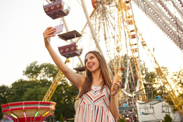 Beautiful long haired positive woman standing over ferris wheel with ice cream cone while making selfie on her mobile phone, being in nice mood and smiling cheerfully