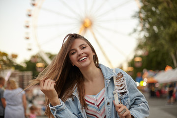 Charming pretty young lady with long brown hair posing over ferris wheel in casual clothes, waving her hair and smiling cheerfully to camera, positive emotions concept