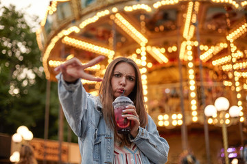 Young beautiful long haired woman grimacing while drinking lemonade, looking to camera and frowning, standing over carousel with raised fingers in victory gesture