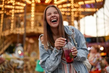 Fotobehang Vrolijke mooie jonge vrouw met bruin haar in casual kleding limonade drinken tijdens het wandelen in pretpark, luid lachen met gesloten ogen en rimpelen © timtimphoto