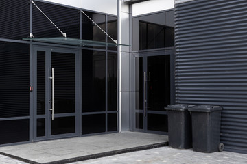 Glass doors - entrance to modern building and two scratched black dustbins against black metal corrugated wall nearby