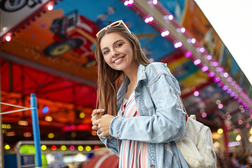 Outdoor photo of positive young pretty woman with long brown hair looking to camera with sincere wide smile, wearing white backpack, trendy jeans coat and light summer dress