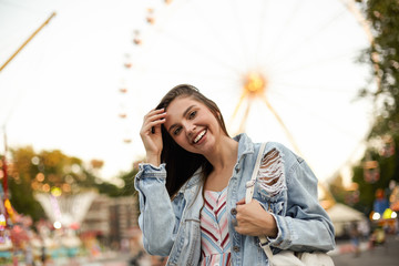 Young beautiful cheerful brunette female in trendy jeans coat standing over ferris wheel in amusement park, looking to camera happily and straightening her hair