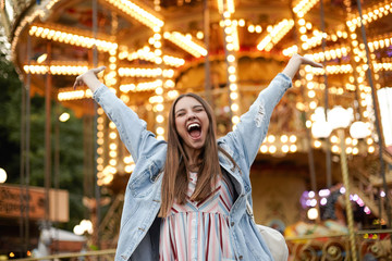 Gelukkig mooie jonge vrouw met lang bruin haar poseren over carrousel in park van attracties, handen gelukkig opsteken en breed lachend met gesloten ogen, casual kleding dragen