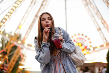 Young attractive long haired female standing over ferris wheel in park of attractions and drinking lemonade with straw, looking to camera and giving wink