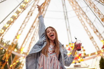 Positive young beautiful brunette woman posing over attractions in amusement park with cup of lemonade in hand, raising palm upwards and smiling broadly to camera