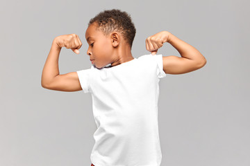 Children, fitness and bodybuilder concept. Studio shot of athletic muscled African American boy in casual t-shirt demonstrating strength by tensing bicep muscles, having proud confident look