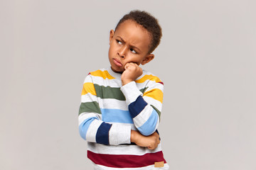 Waist up shot of moody grumpy African schoolboy in sweater holding hand on his face, pouting lips...
