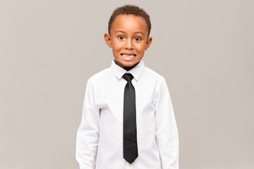 Shy or scared emotional African schoolboy in white shirt and black tie baring his teeth. Portrait of funny dark skinned child in formal clothes grimacing against blank studio wall background