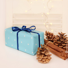gift in a cardboard box is tied with a blue satin ribbon, beautiful pine cones and a bunch of cinnamon sticks on a wooden table on a light festive background with luminous lights of garlands