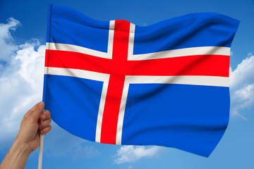 hand holds iceland color flag on sky background with clouds on luxurious satin texture, silk with waves, closeup, copy space