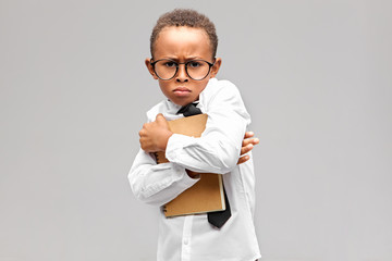 Picture of angry frowning dark skinned straight A pupil in glasses and white uniform holding tight...