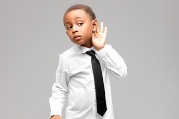 Horizontal shot of curious Afro American schoolboy listening, making hand gesture to his ear. Dark...