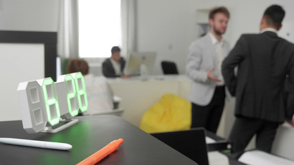 Clock is on the shelf, on the backgorund there are office workers.
