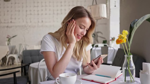 Charming happy young blonde woman smiling and using smartphone while sitting in cafe
