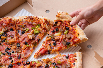 Child hand taking slice of delicious Italian pizza with bacon, olives, sausages and mushrooms. Top view. Fresh tasty pizza in cardboard box - close-up.