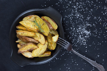 Top view of rustic roasted potatoes with rosemary branch on black plate on black background