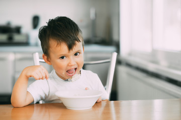 A little boy in the kitchen eating oatmeal from a white plate