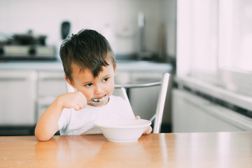 A little boy in the kitchen eating oatmeal from a white plate