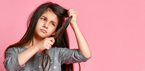 little brunette girl with long hair holds a strand of her hair and looks at it. Hair care and...
