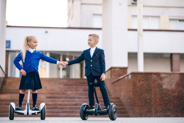 Fototapeta na wymiar Two young schoolchildren riding on gyroscooters. Kids go to school for gyroscooters. Blonde kids. Back to school. Modern schoolchildren. Kids in school uniform. Flags of foreign countries.