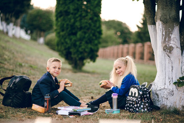 Cute kids having lunch at school break sitting on lawn near school near big tree.  Back to school.