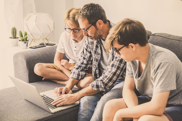 Family sitting on sofà at home working with laptop. Father helping a young brothers making homework Wireless technology allow to stay connected everywhere Beautiful familiar scene of dad and two sons