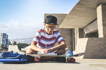 Trendy student doing homework out of the college sitting on a concrete wall. Young boy writing on...