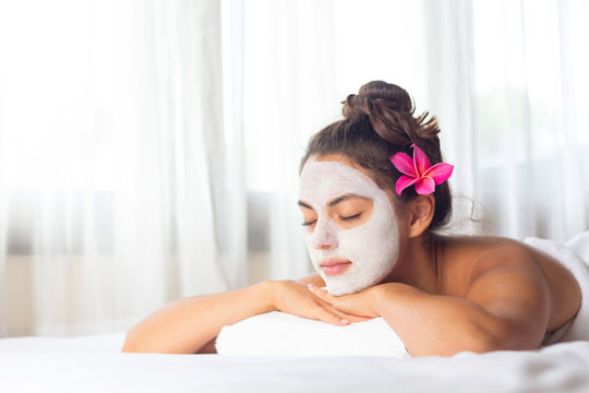 Nice relaxing young woman with white mask on face lying down on towel on bench spa with flower on hair. Smiling on camera.