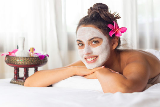 Nice relaxing young woman with white mask on face lying down on towel on bench spa with flower on hair. Smiling on camera.