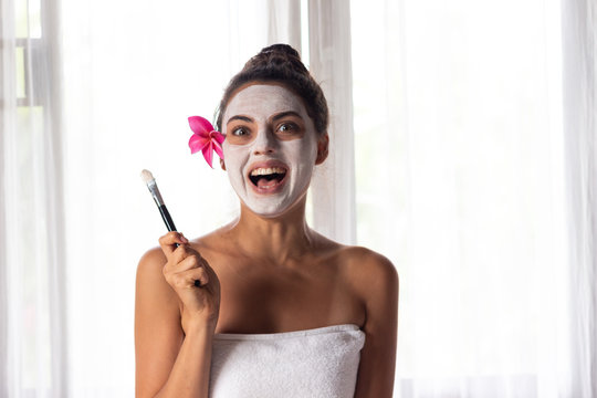 Nice relaxing young woman with white mask on face lying down on towel on bench spa with flower on hair. Smiling on camera.