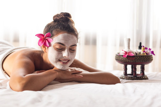 Nice relaxing young woman with white mask on face lying down on towel on bench spa with flower on hair. Smiling on camera.