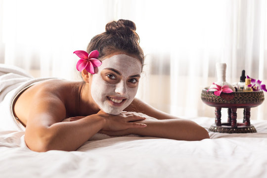 Nice relaxing young woman with white mask on face lying down on towel on bench spa with flower on hair. Smiling on camera.