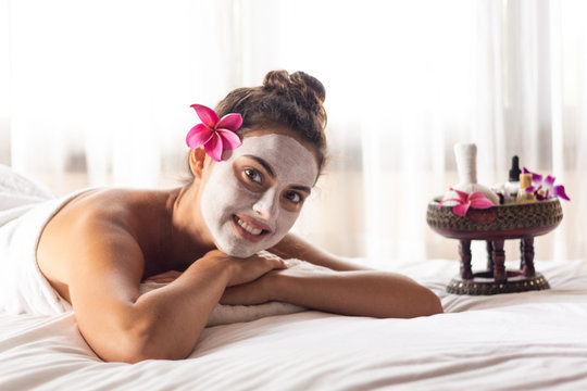 Nice relaxing young woman with white mask on face lying down on towel on bench spa with flower on hair. Smiling on camera.