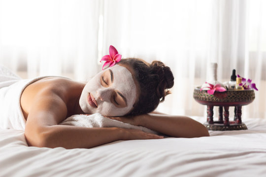 Nice relaxing young woman with white mask on face lying down on towel on bench spa with flower on hair. Smiling on camera.