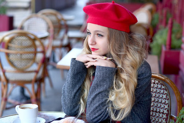 blonde girl in a red beret and in a gray coat with long curls sits thoughtfully at a table in a cafe