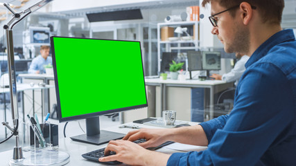 Over the Shoulder Shot of Engineer Working with Green Screen Mock-up Template on Desktop Computer. In the Background Engineering Facility Specialising on Industrial Design