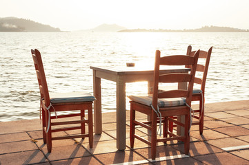 Fototapeta na wymiar Beach cafe with sea view at sunset, empty table, three chairs, vacant for guests. Simple, authentic wooden furniture. Beautiful summer holiday concept. Horizon with misty mountains on distant island