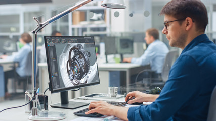 Over the Shoulder Shot of Engineer Working with CAD Software on Desktop Computer, Screen Shows Technical Details and Drawings. In the Background Engineering Facility Specialising on Industrial Design