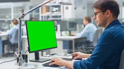 Over the Shoulder Shot of Engineer Working with Green Mock-up Screen Desktop Computer. In the Background Engineering Facility with Specialists Working on Blueprints and Drawings with Industrial Design