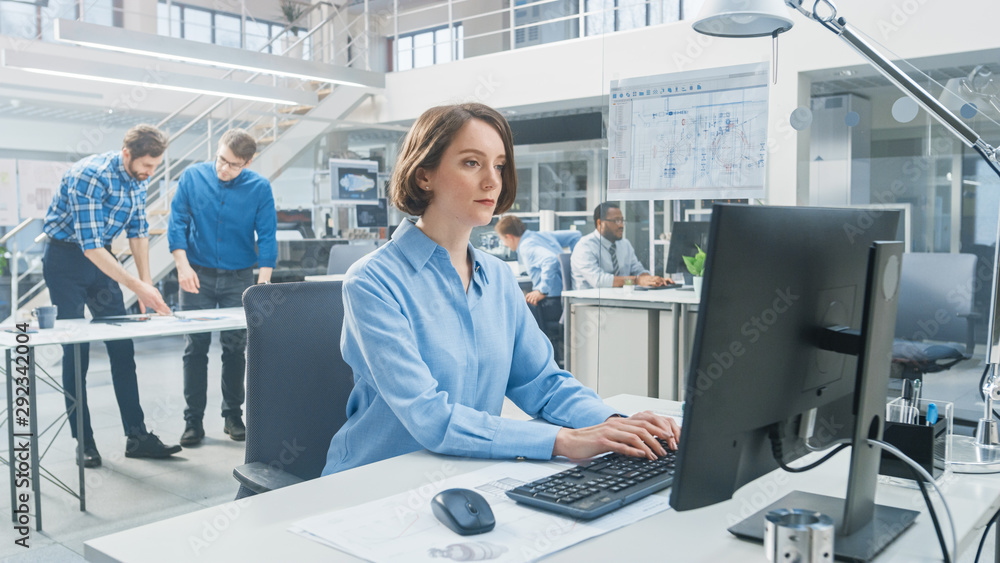 Poster In the Industrial Engineering Facility: Portrait of the Smart and Beautiful Female Engineer Working on Desktop Computer. In the Background Specialists and Technicians Working with Drafts and Drawings