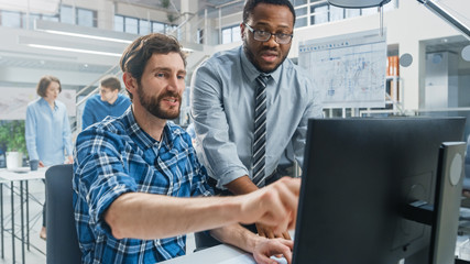 In the Industrial Engineering Facility: Portrait of the Handsome Male Engineer Working on Desktop Computer, Chief Engineer Stands Beside Overseeing Project. They Succeed